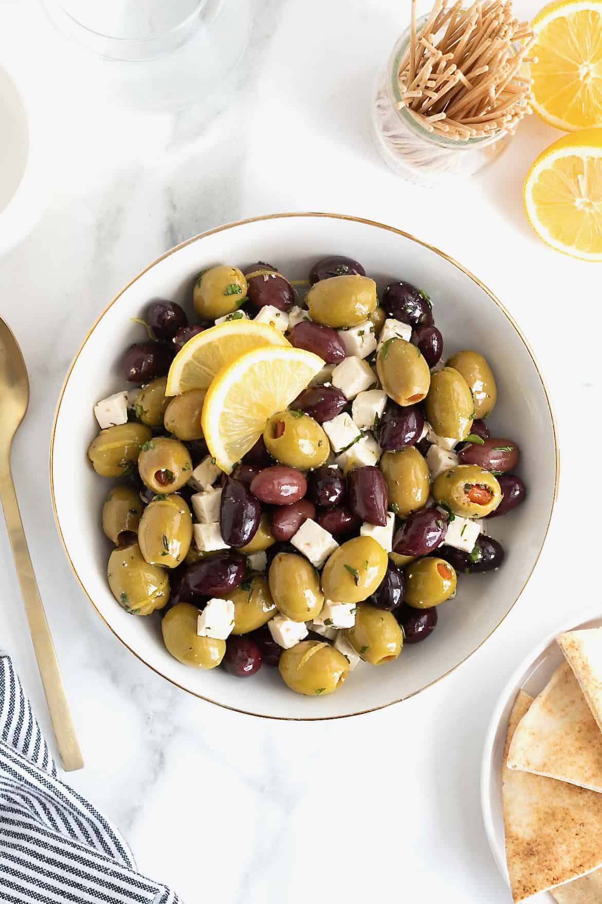 A gold rimmed white serving bowl filled with green and purple olives and small cubes of feta.