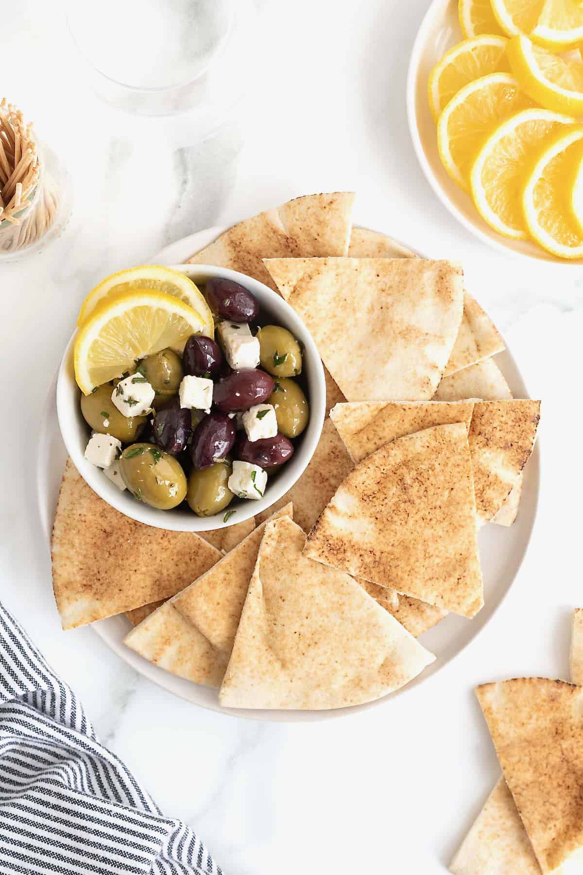A bowl of marinated olives and feta surrounded by pita chips on a white serving platter.