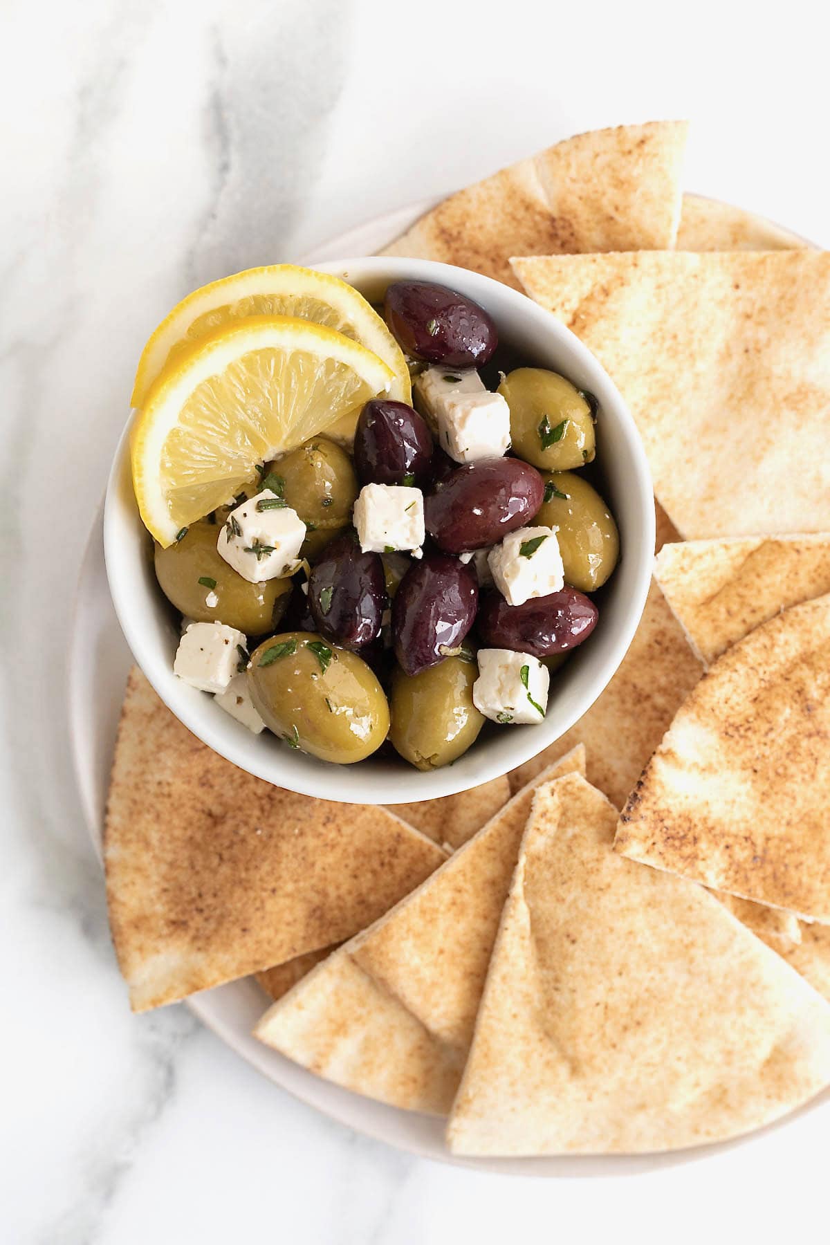 A bowl of marinated olives and feta surrounded by pita chips on a white serving platter.