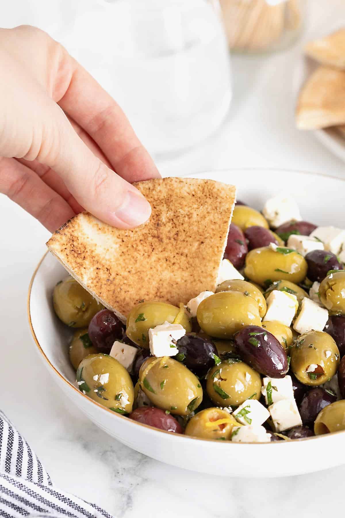 Marinated olives and feta being scooped onto a pita chip from a gold-rimmed white bowl.