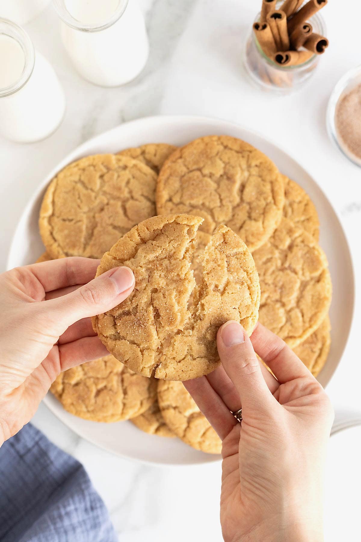 Two hands breaking a snickerdoodle cookie in half over a plate of cookies.