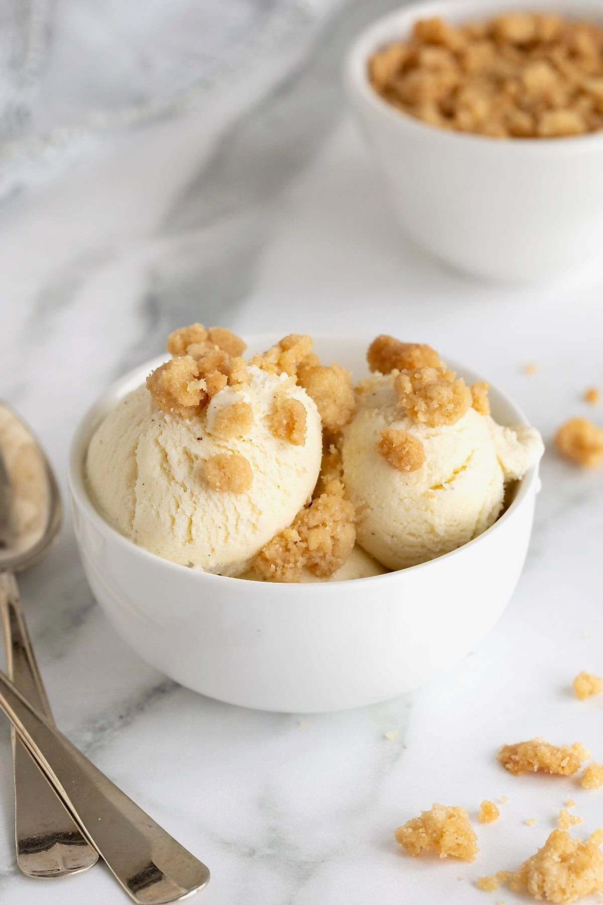 Vanilla ice cream topped with simple streusel in a white bowl next to a metal spoon on a white counter.