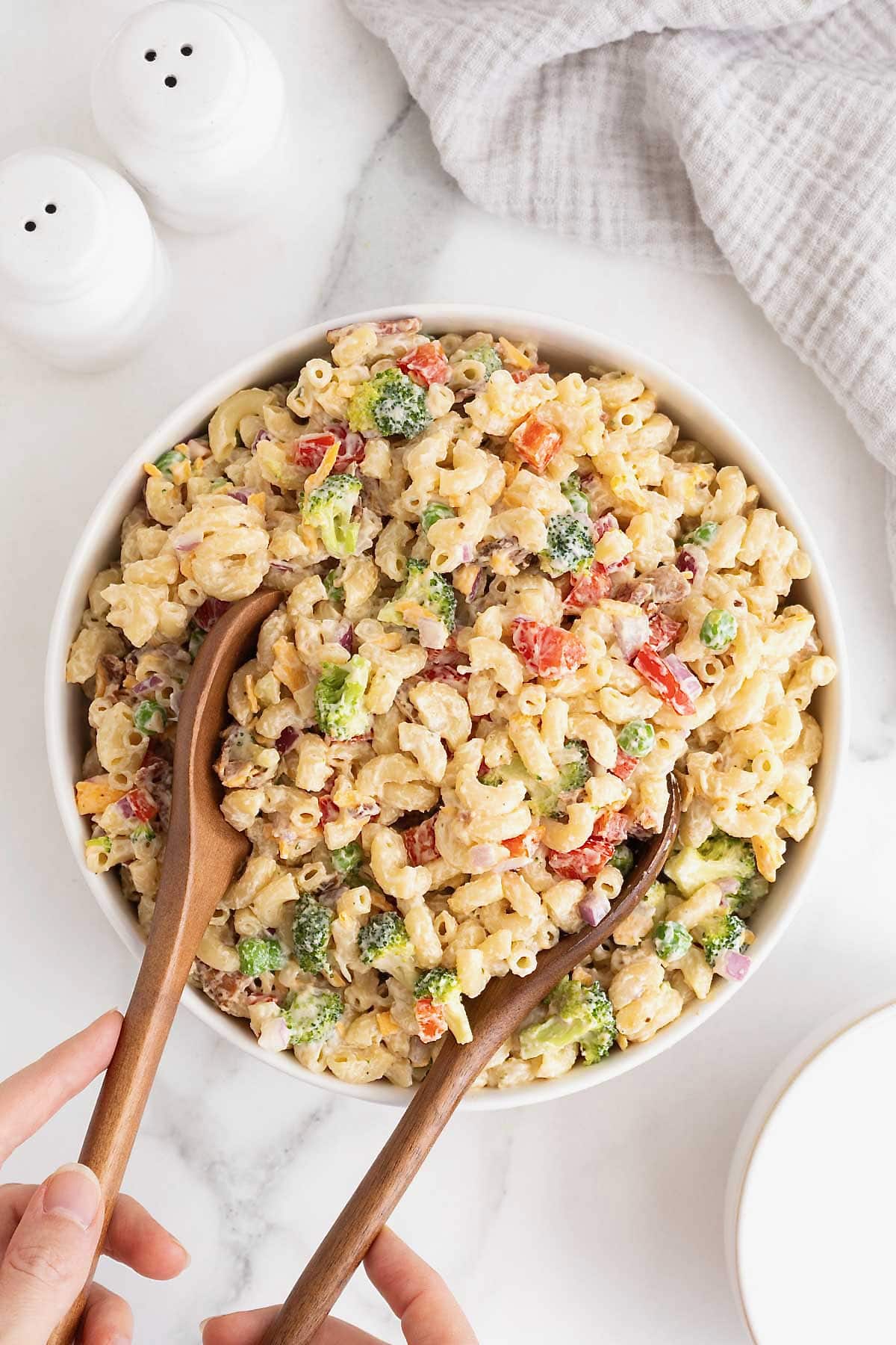 Two wooden serving spoons in a large white serving bowl of macaroni salad with sweet peas, red onion and cheese.