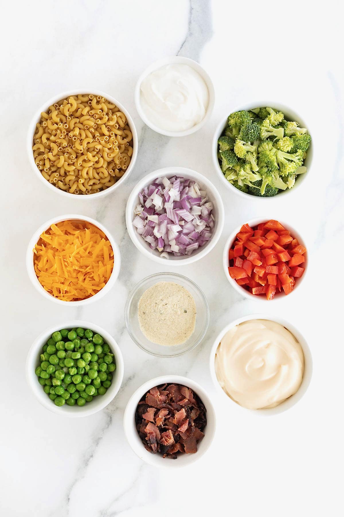 Ingredients to make bacon ranch macaroni salad in small white dishes on a white marble counter.