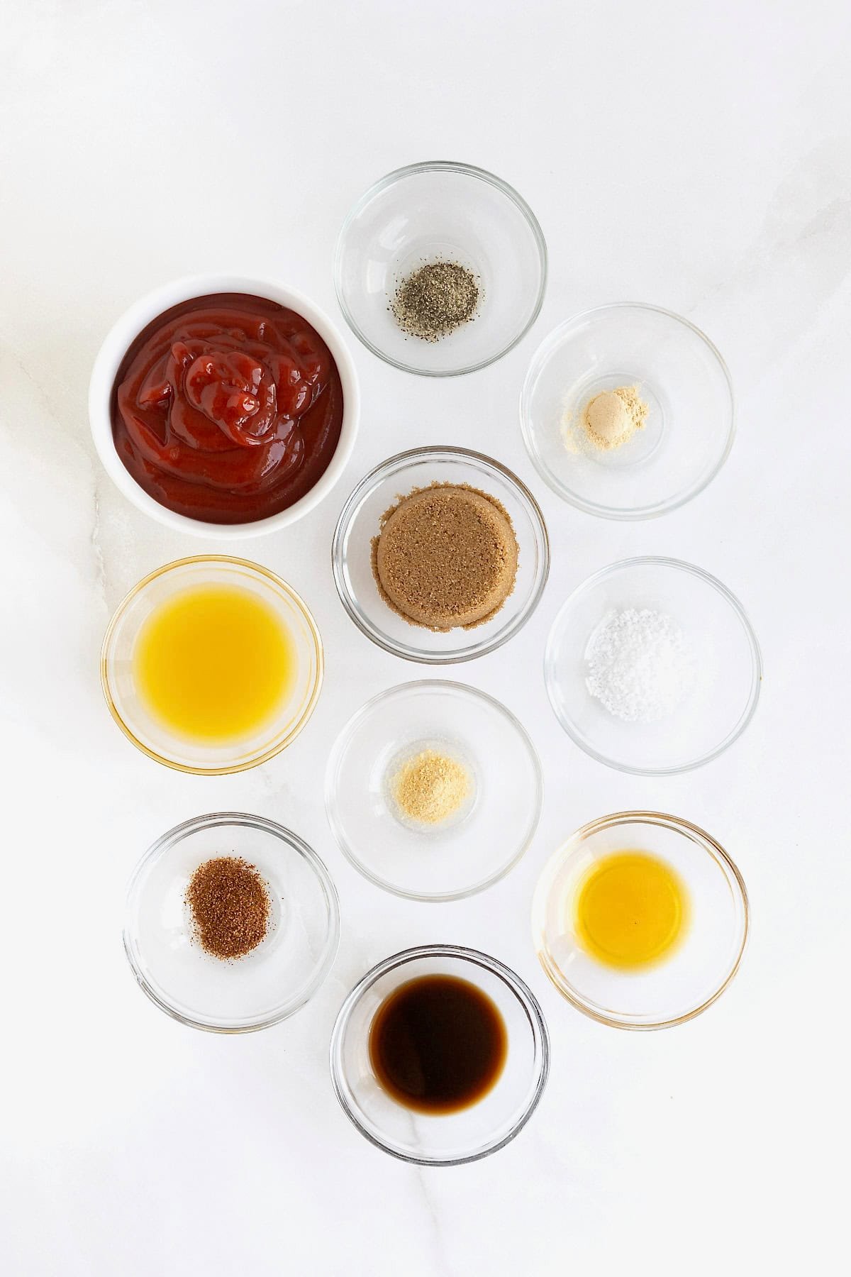 Ingredients to make homemade barbecue sauce in small glass dishes on a white marble counter.