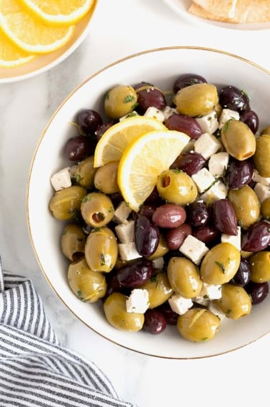 A gold rimmed white serving bowl filled with green and purple olives and small cubes of feta.