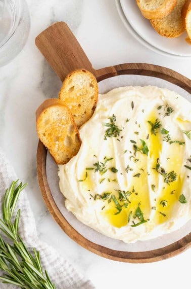 A wooden serving plate of whipped brie drizzled with olive oil. Two pieces of crostini are stuck in the brie.