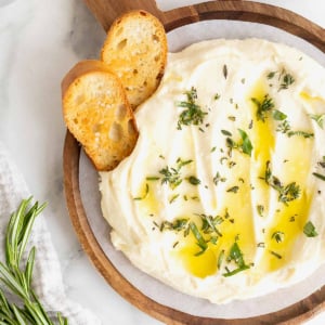 A wooden serving plate of whipped brie drizzled with olive oil. Two pieces of crostini are stuck in the brie.
