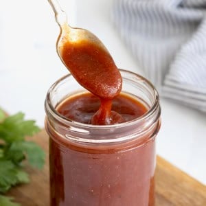 A spoon being dipped into a wide mouth mason jar of barbecue sauce on a wooden cutting board.
