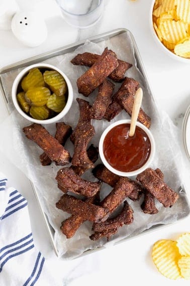 A parchment lined aluminum baking sheet of baby back ribs. There is a small white dish of pickles at the top left and a small white dish of barbecue sauce in the center.