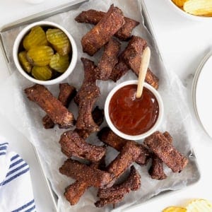 A parchment lined aluminum baking sheet of baby back ribs. There is a small white dish of pickles at the top left and a small white dish of barbecue sauce in the center.