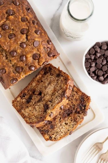 A loaf of chocolate chip banana bread on a marble cutting board. A small white dish of chocolate chips sits to the right side.