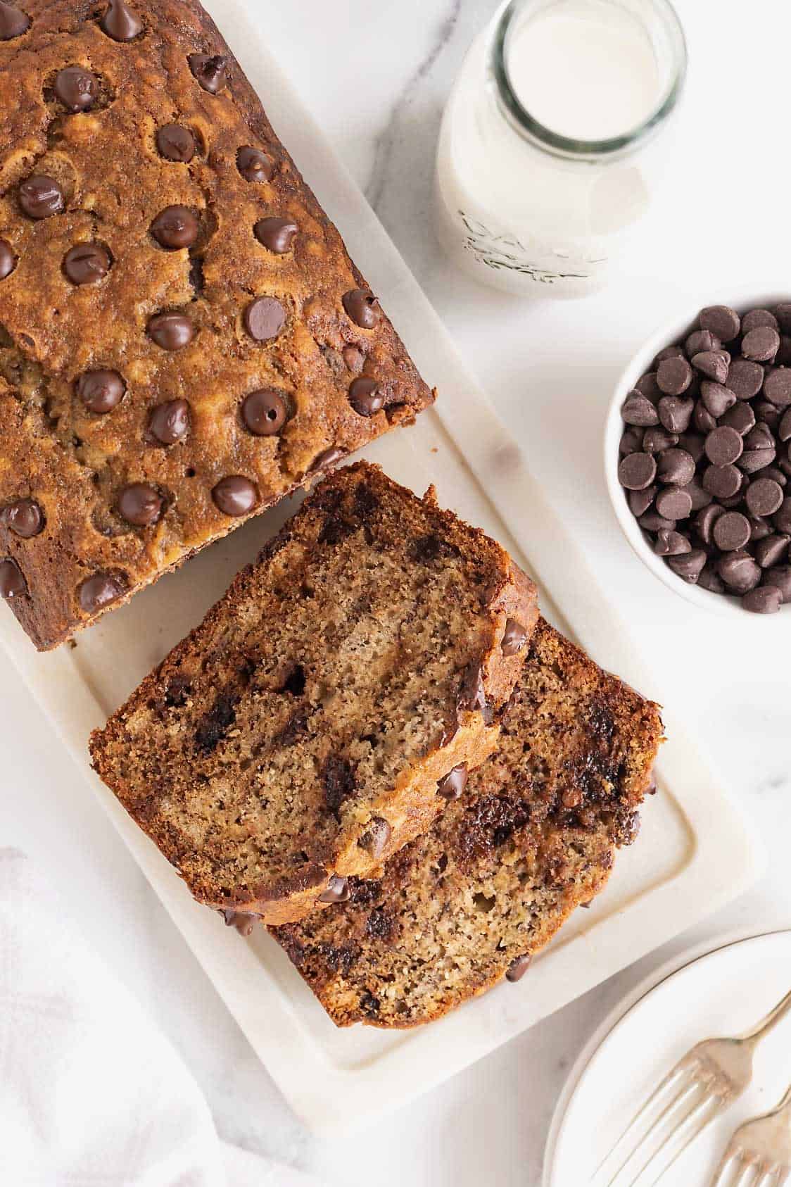 A loaf of chocolate chip banana bread on a marble cutting board. A small white dish of chocolate chips sits to the right side.