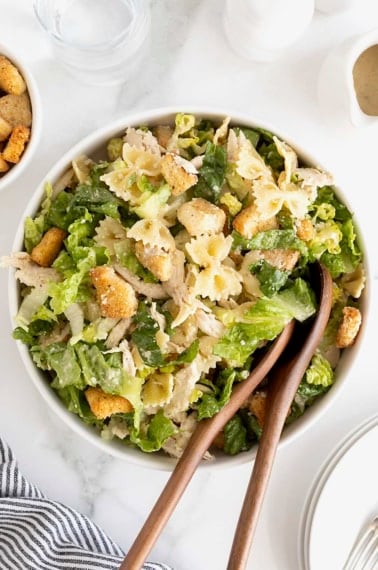 A large white salad bowl of caesar pasta salad with chicken. There are two wooden serving spoons in the bowl.