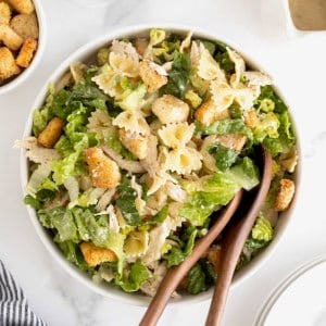 A large white salad bowl of caesar pasta salad with chicken. There are two wooden serving spoons in the bowl.