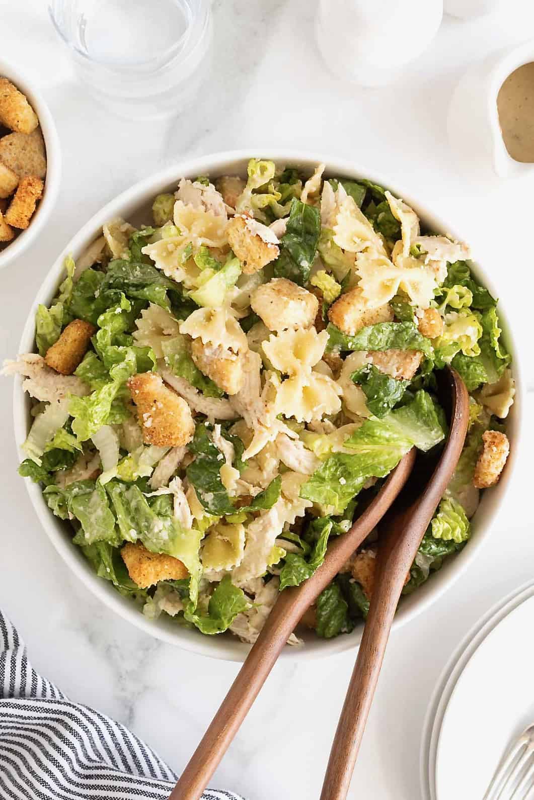 A large white salad bowl of caesar pasta salad with chicken. There are two wooden serving spoons in the bowl.