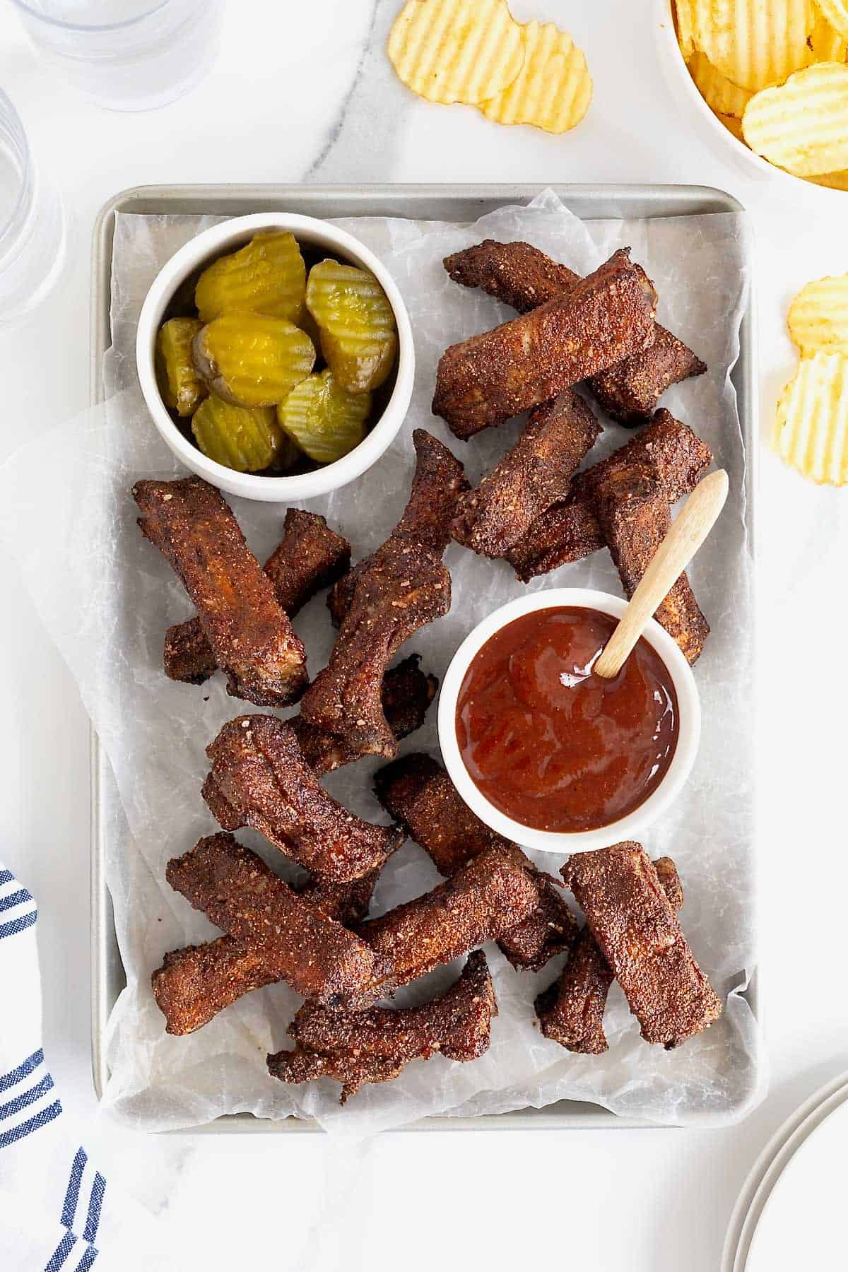 A parchment lined aluminum baking sheet of baby back ribs. There is a small white dish of pickles at the top left and a small white dish of barbecue sauce in the center.