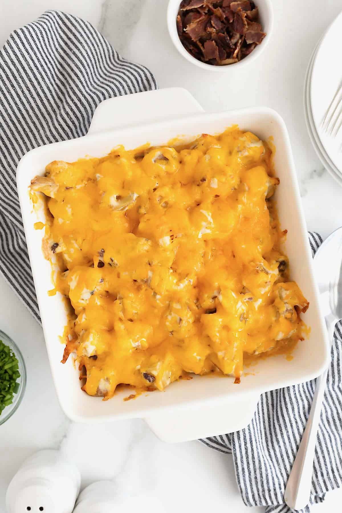 A square white baking dish of potato casserole topped with bacon bits and melted cheese.
