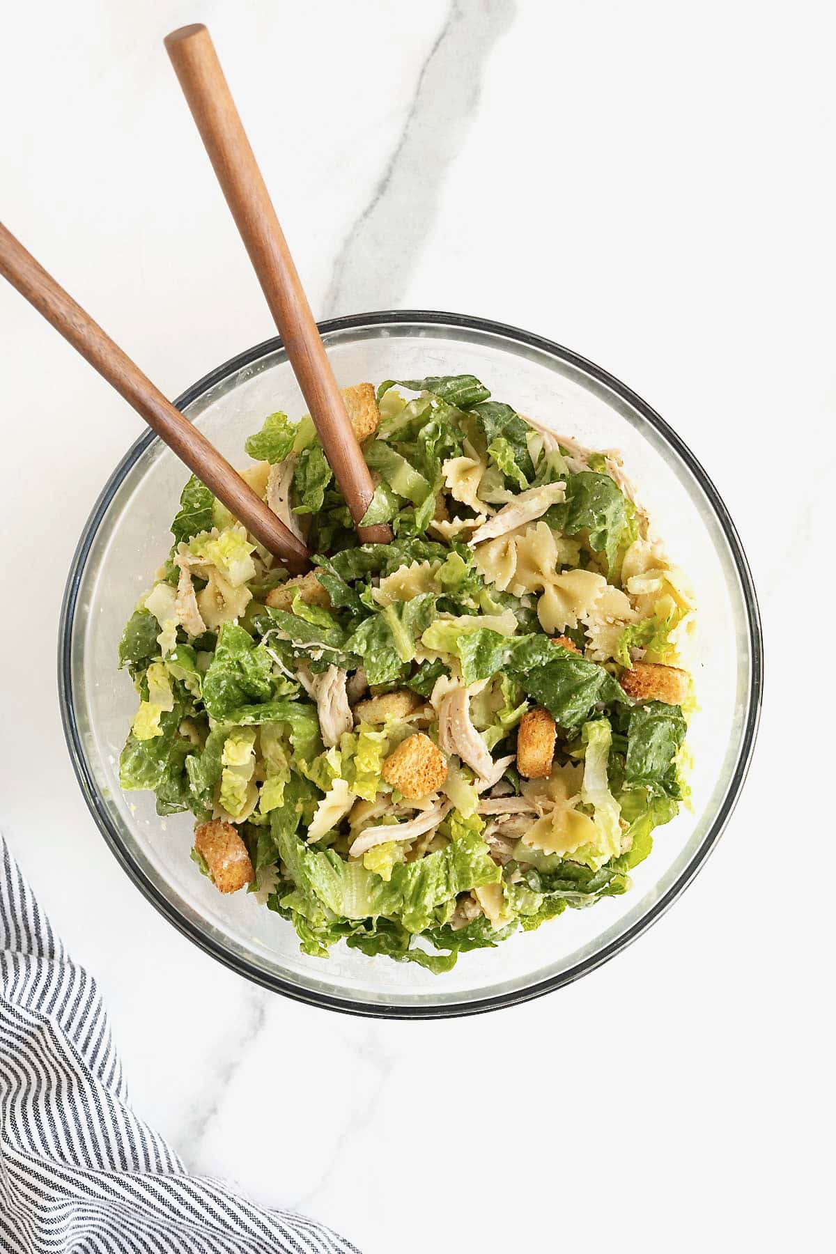 A large glass mixing bowl of caesar pasta salad with chicken. There are two wooden serving spoons in the bowl.