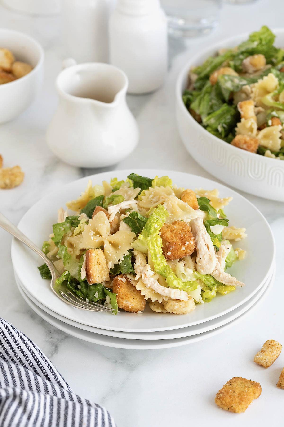 A stack of three white salad plates with a serving of chicken caesar pasta salad on the top plate. A fork is resting on the plate.