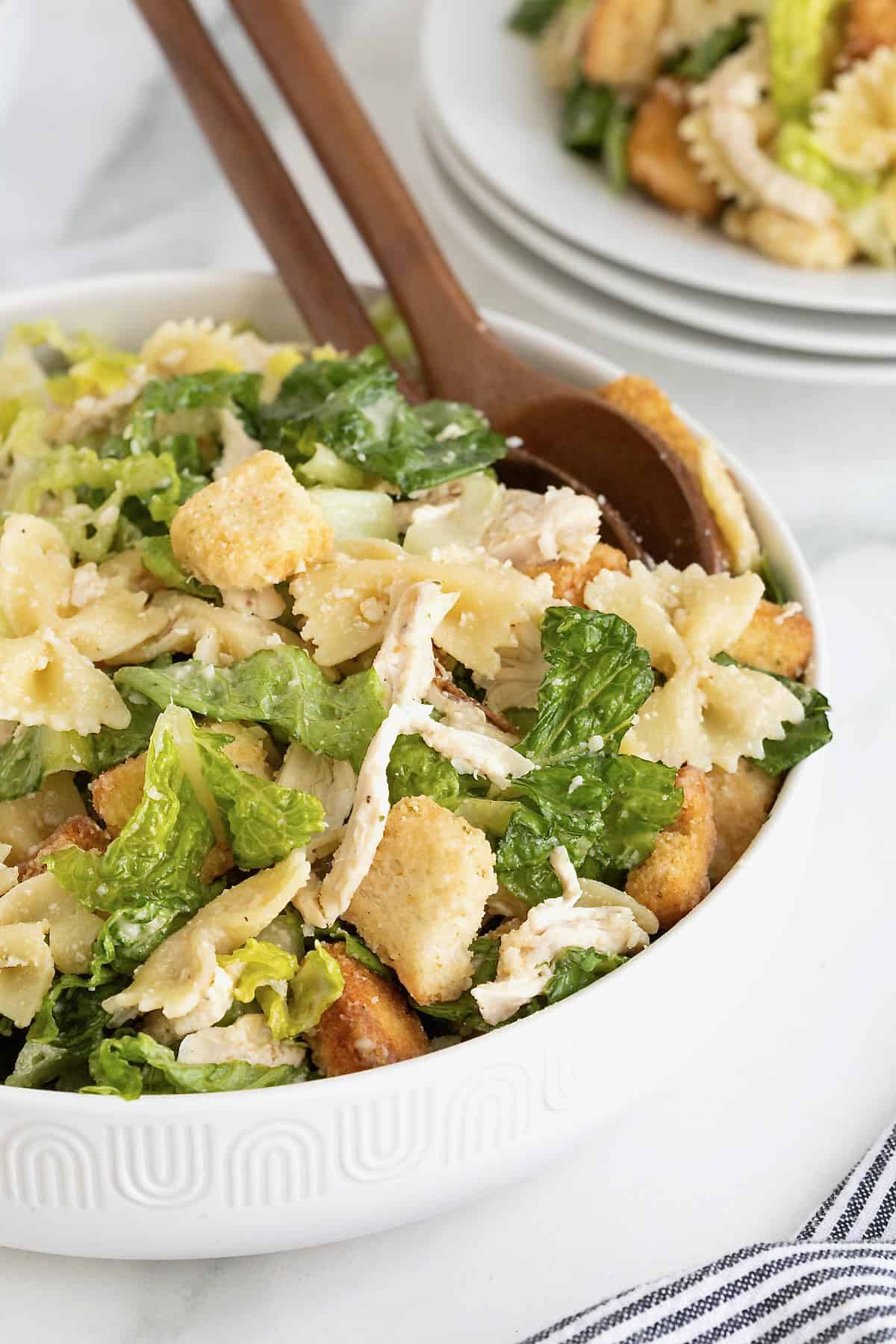 A large white salad bowl of caesar pasta salad with chicken. There are two wooden serving spoons in the bowl.