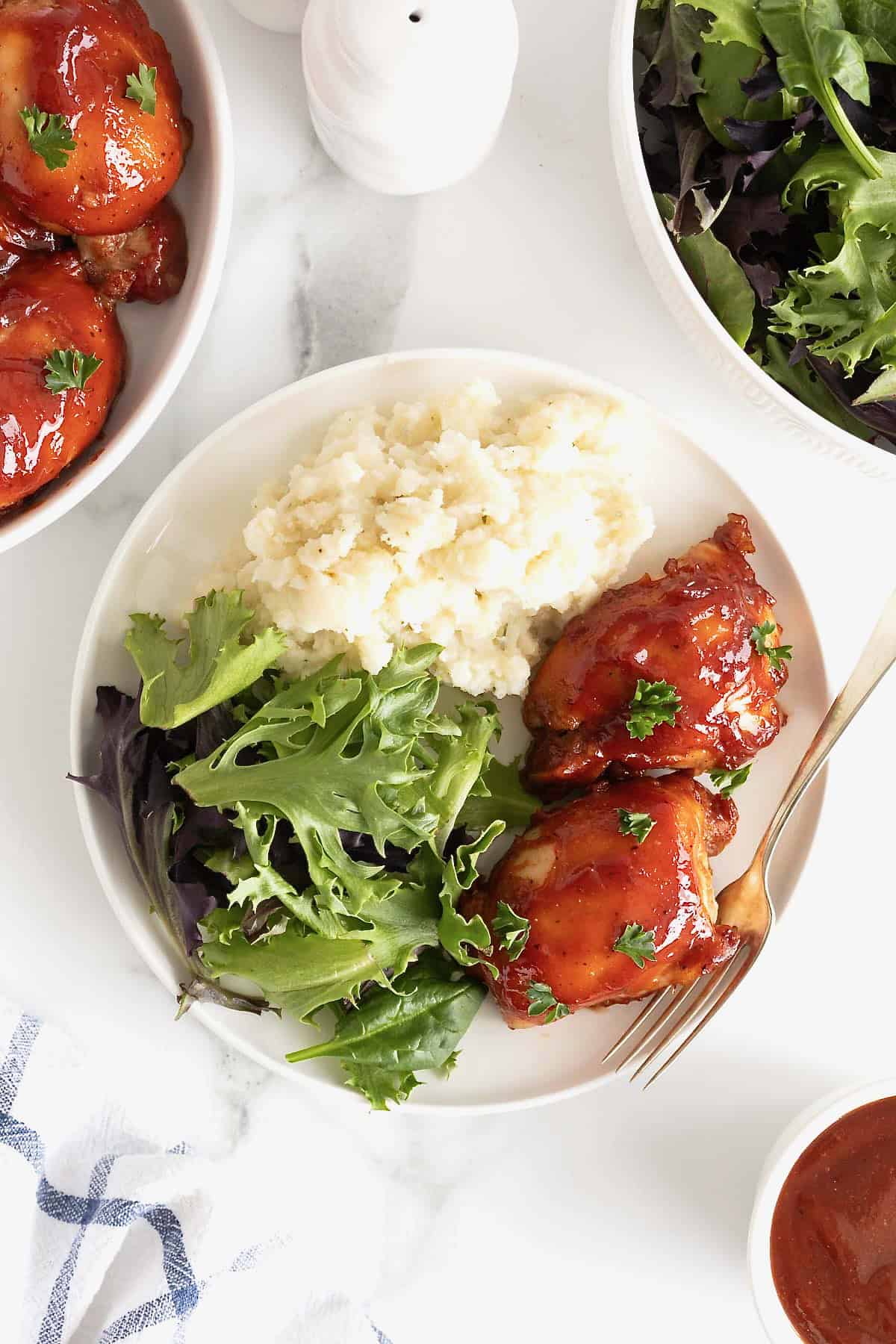A white dinner plate with two bbq chicken thighs, a green salad, and mashed potatoes.