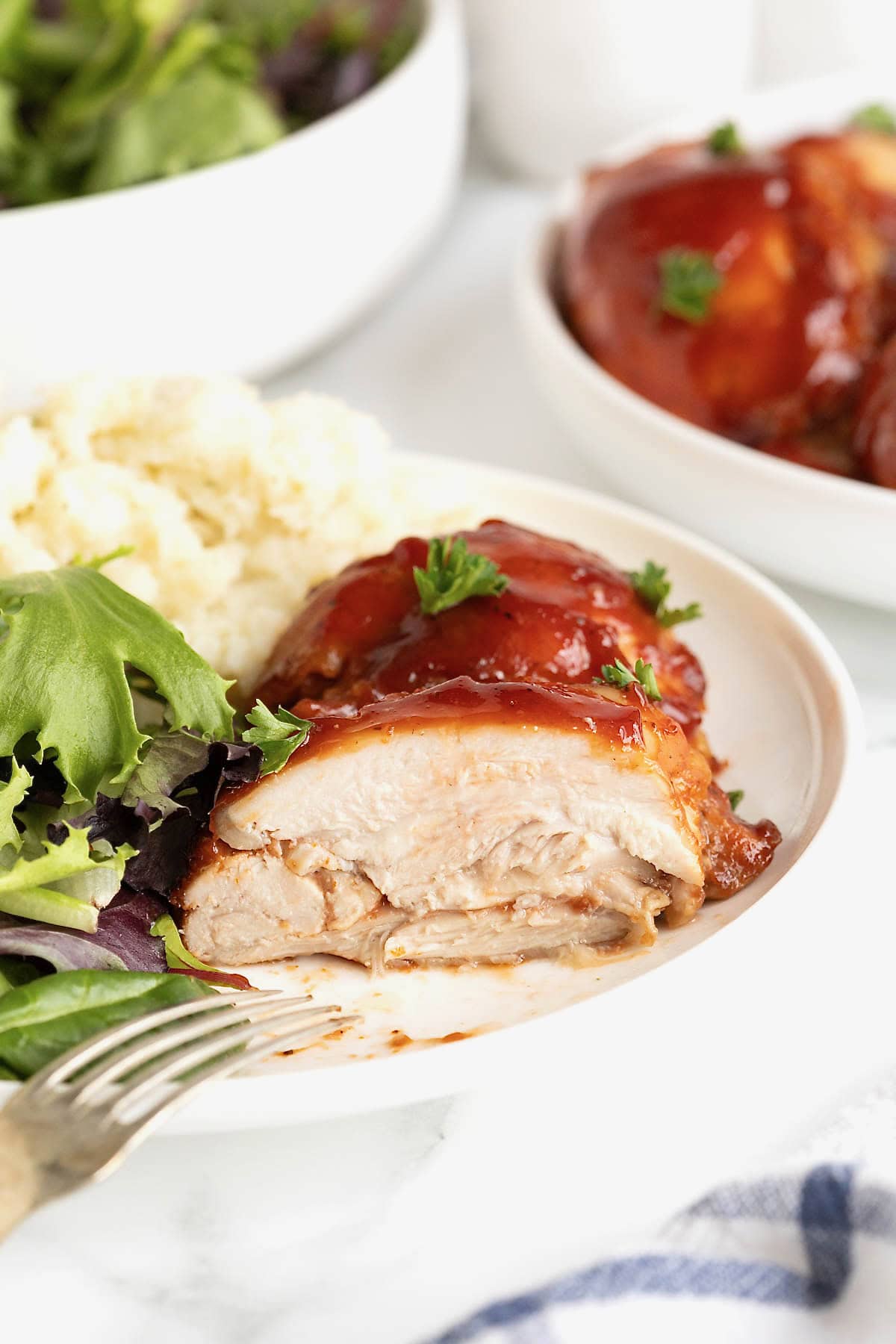 A piece of barbecue chicken on a white dinner plate next to mashed potatoes and a green salad.