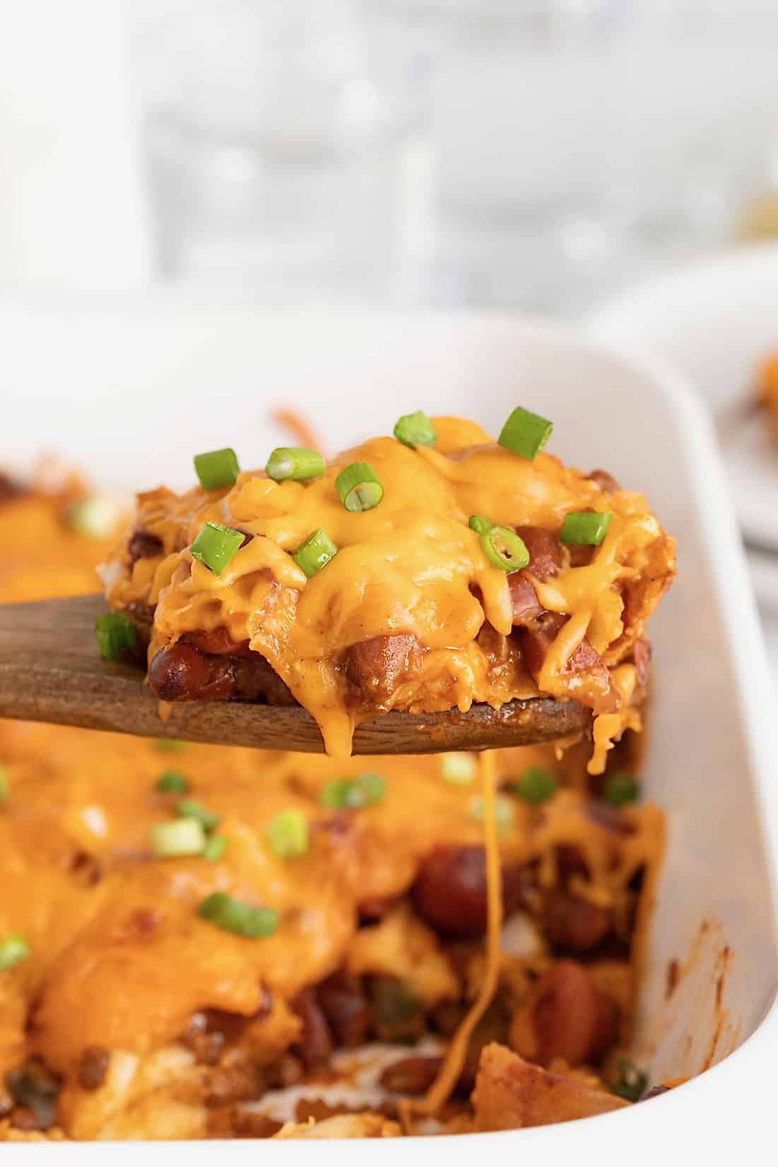 A wooden spoon lifting a serving of chili cheese dog casserole from a white casserole dish.