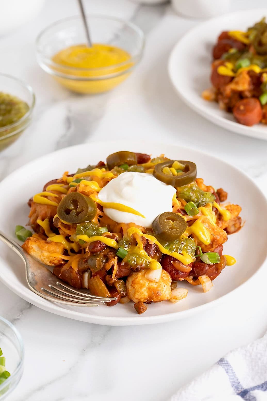 A serving of chili cheese dog casserole topped with a drizzle of mustard, a dollop of sour cream, and four jalapeño slices on a white plate.