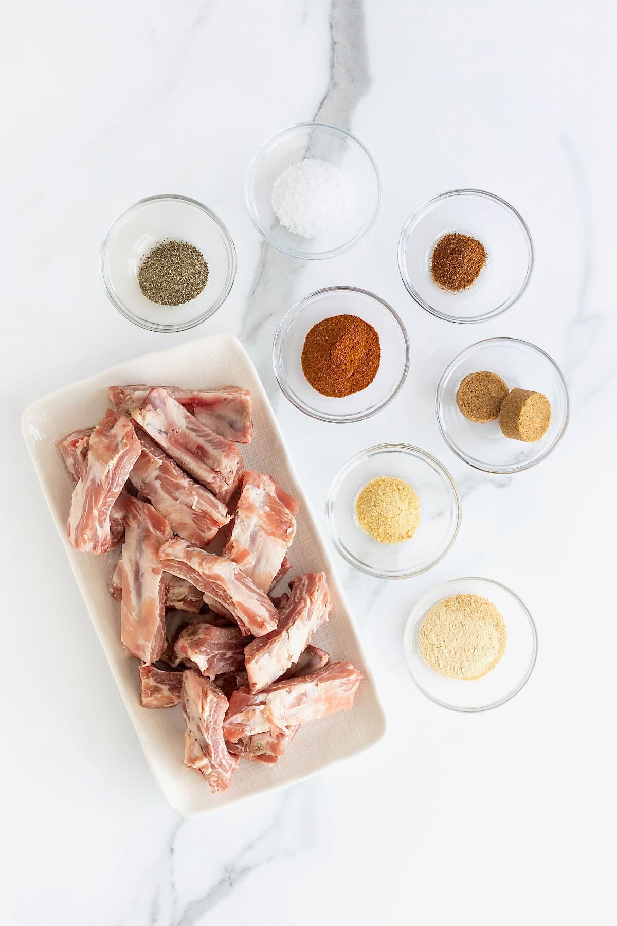Ingredients to make baked baby back ribs with a dry rub. The ingredients are in small glass dishes on a white marble counter.