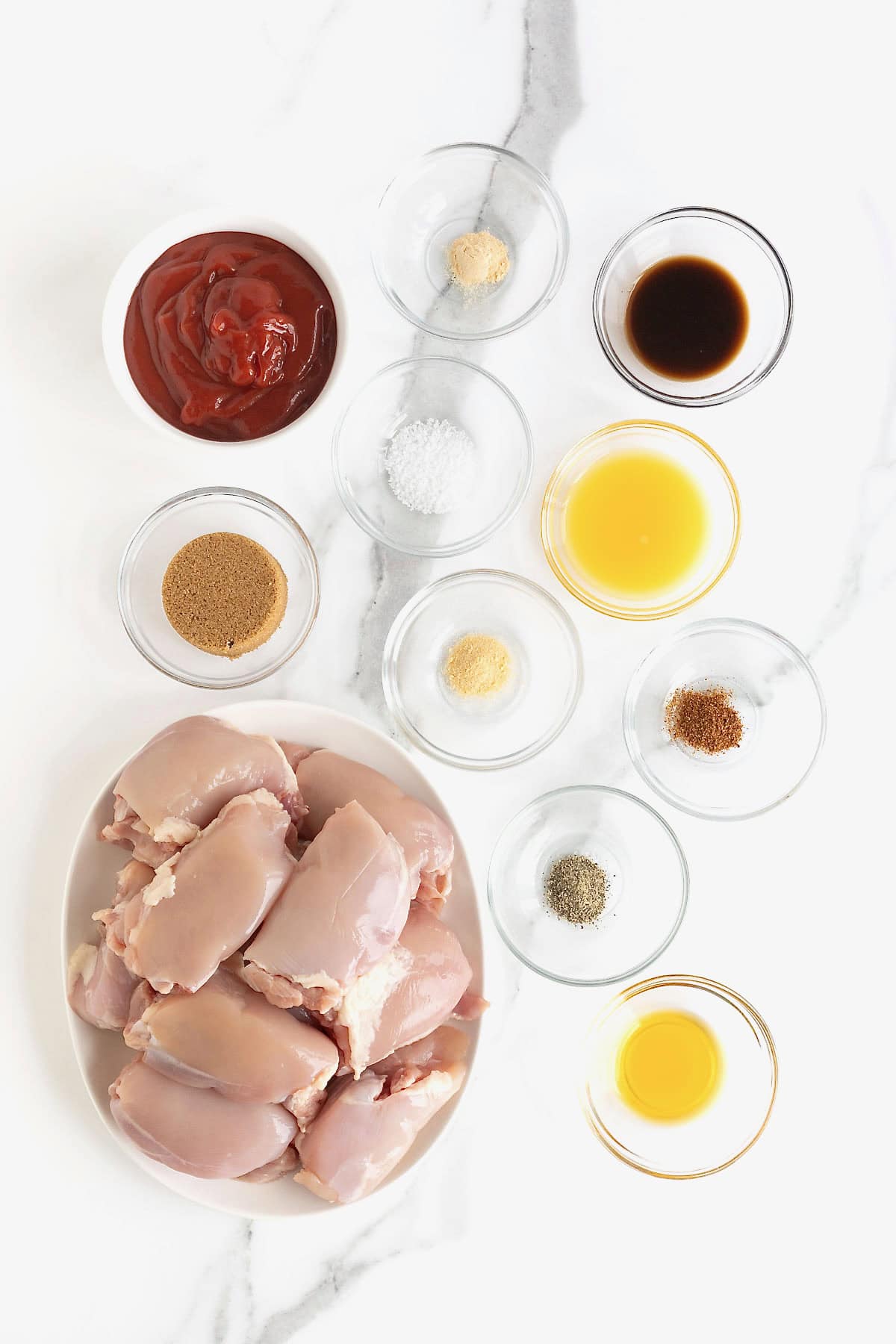 Ingredients to make barbecue chicken thighs in glass dishes on a white marble counter.