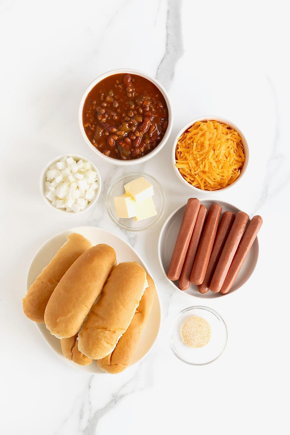 Ingredients to make Chili Cheese Dog Casserole in white dishes on a white marble counter.