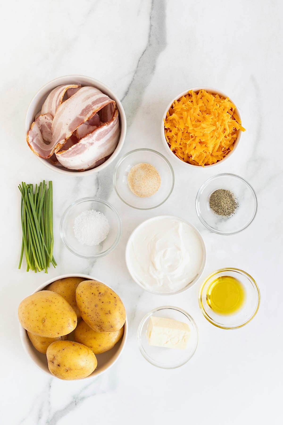 Ingredients to make Twice Baked Potato Casserole ins mall glass dishes on a white marble counter.