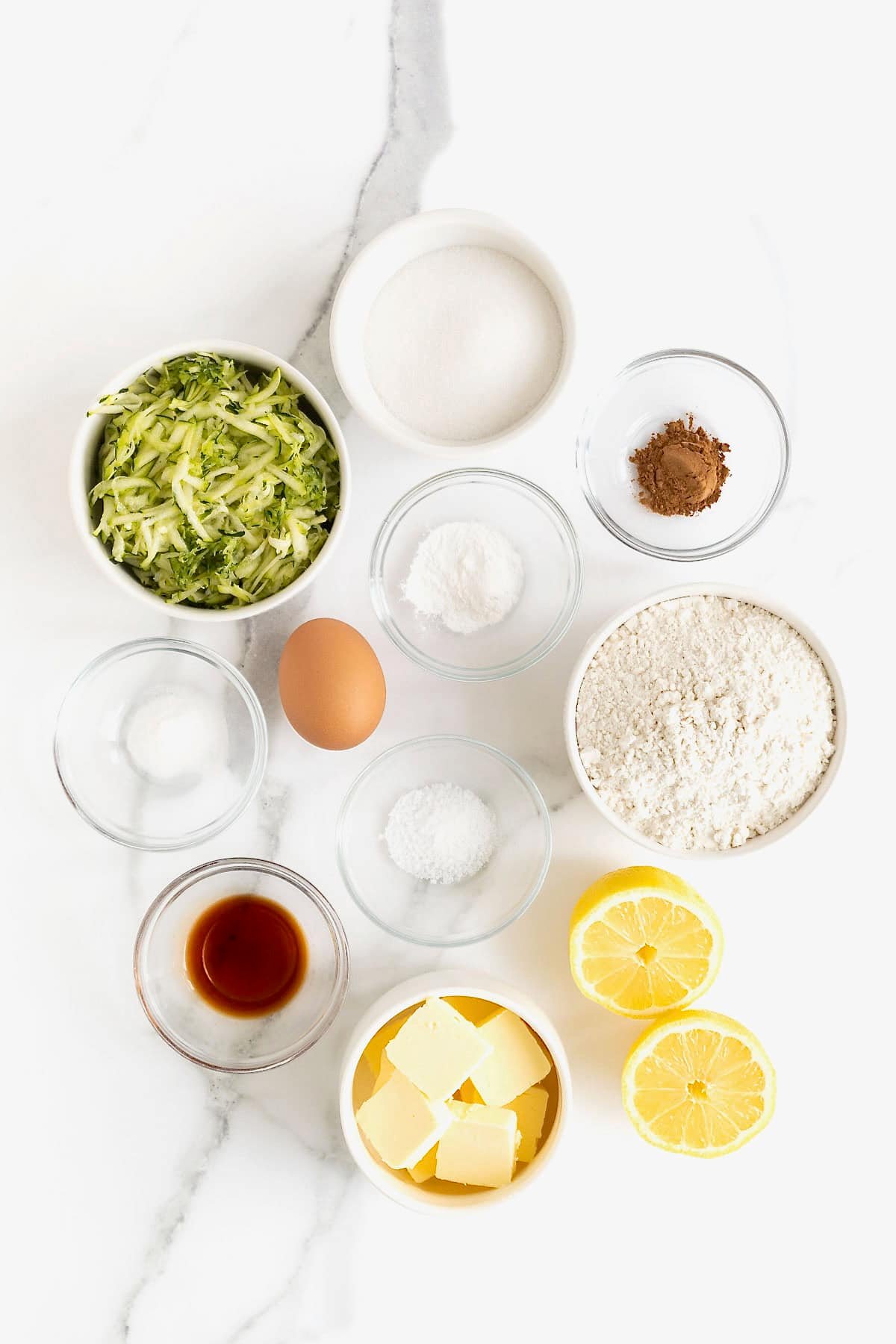 Ingredients to make zucchini muffins in small glass dishes on a white marble counter.