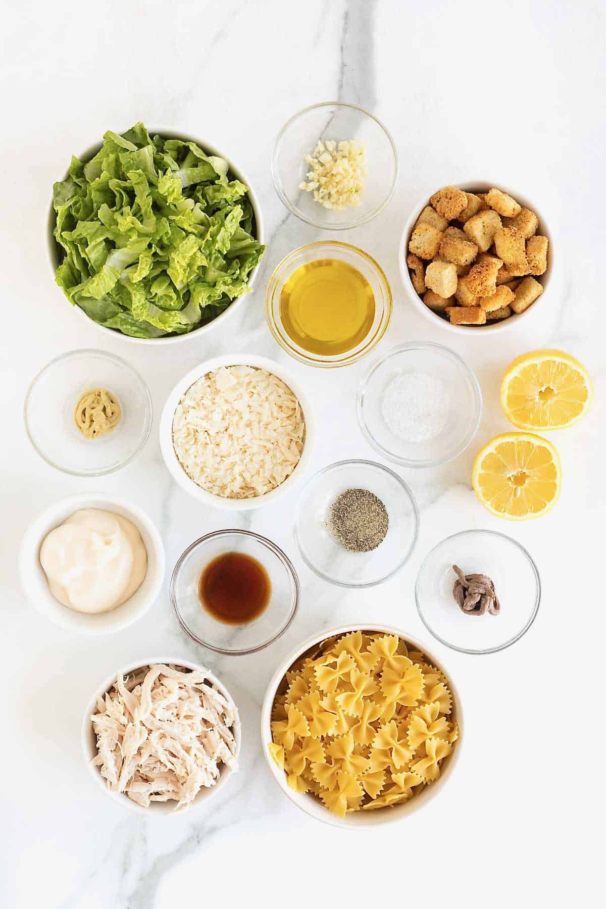 Ingredients to make chicken Caesar pasta salad in small glass dishes on a white marble counter.