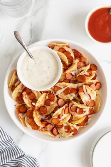 A large rimmed white serving plate piled with pizza taco bites. There is a small white dish of ranch dipping sauce.