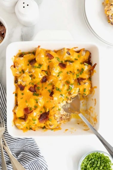 A square white baking dish of potato casserole topped with bacon bits and melted cheese. A serving has been removed and there is a metal spoon in the dish.