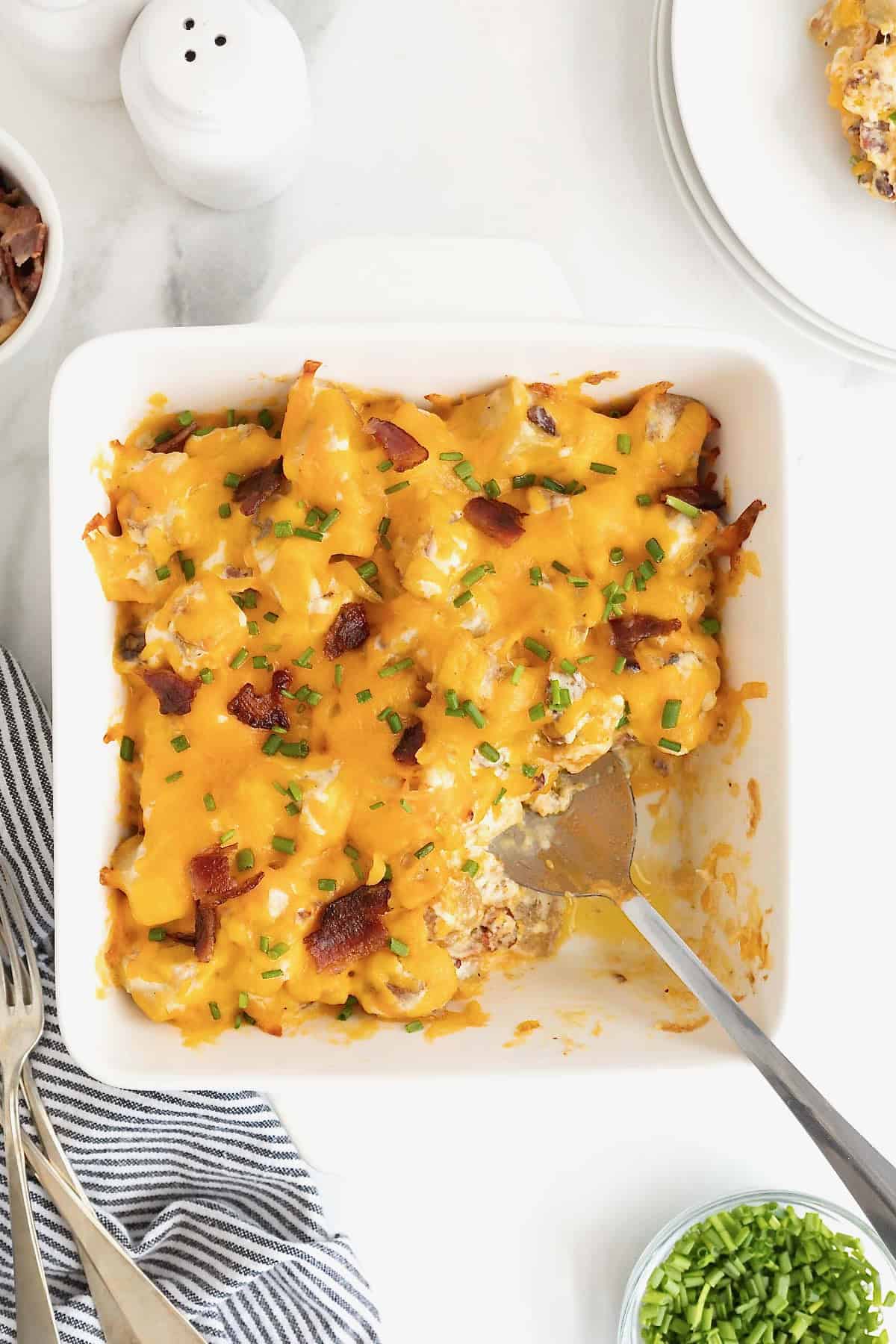 A square white baking dish of potato casserole topped with bacon bits and melted cheese. A serving has been removed and there is a metal spoon in the dish.