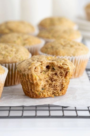 A bite taken out of an unwrapped zucchini muffin on a parchment lined cooling rack. Behind the muffin are six more muffins in paper wrappers.