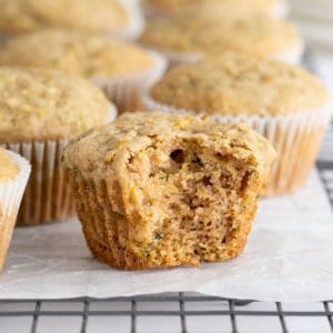 A bite taken out of an unwrapped zucchini muffin on a parchment lined cooling rack. Behind the muffin are six more muffins in paper wrappers.