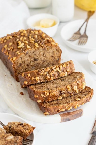 A loaf of banana nut bread on a white cutting board. Half of the loaf is cut into three thick slices.