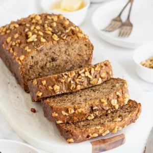 A loaf of banana nut bread on a white cutting board. Half of the loaf is cut into three thick slices.