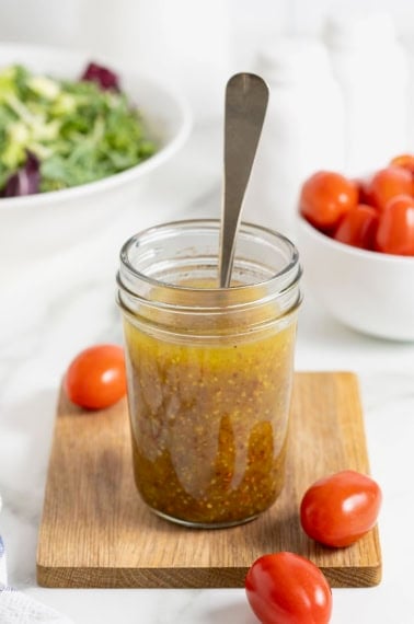 A mason jar of salad dressing with a spoon sticking out of it on a wooden cutting board.