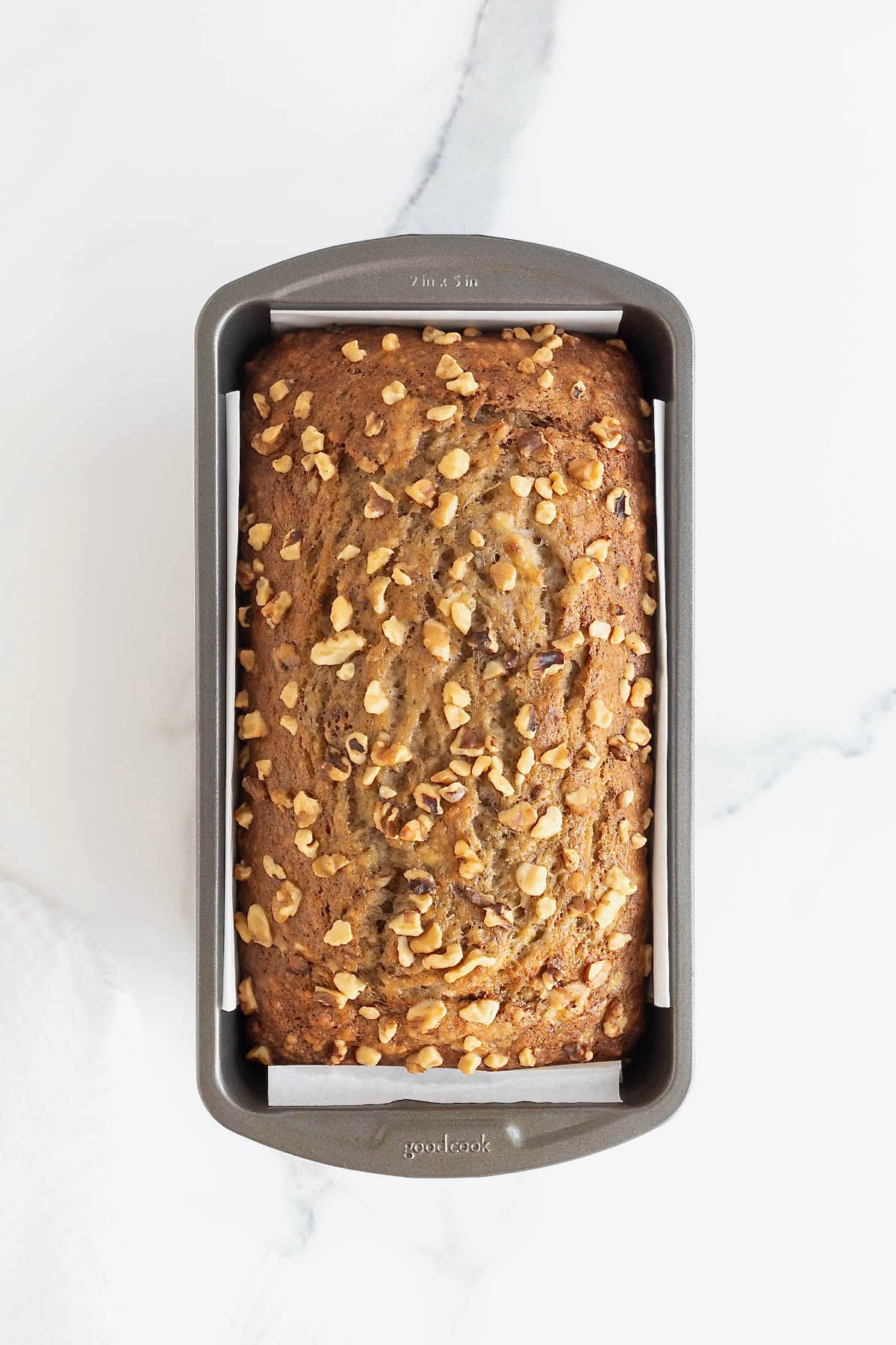 A baked loaf of banana nut bread in a dark aluminum loaf pan on a white marble counter.