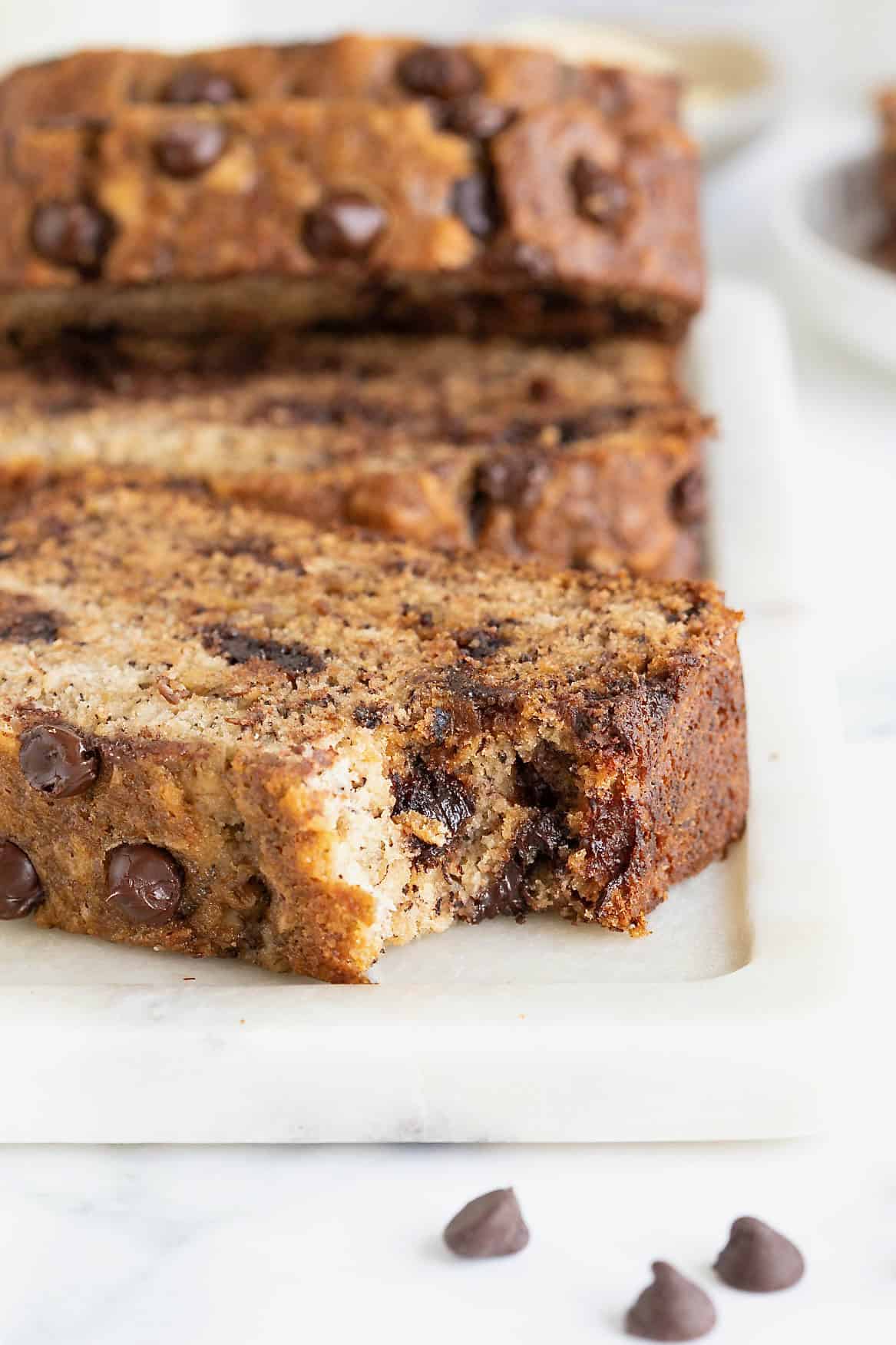 A slice of brown butter banana bread with chocolate chips on a white cutting board. There is a bite taken out of the bread.