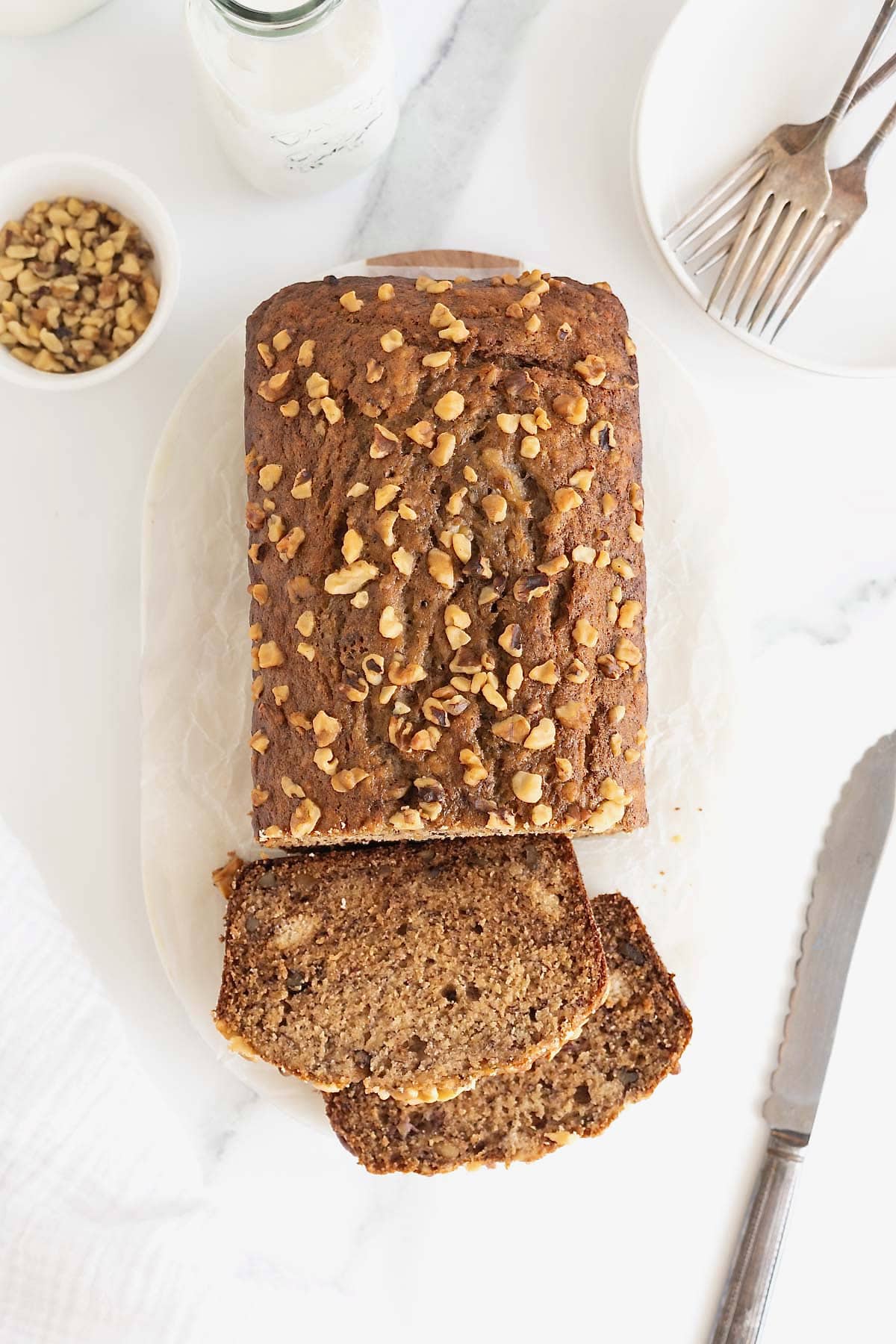 A loaf of banana nut bread on a white cutting board. One quarter of the loaf is cut into two thick slices.