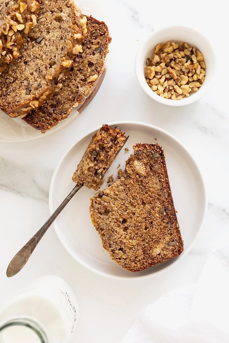 A slice of banana nut bread on a rimmed white plate with a fork next to it.