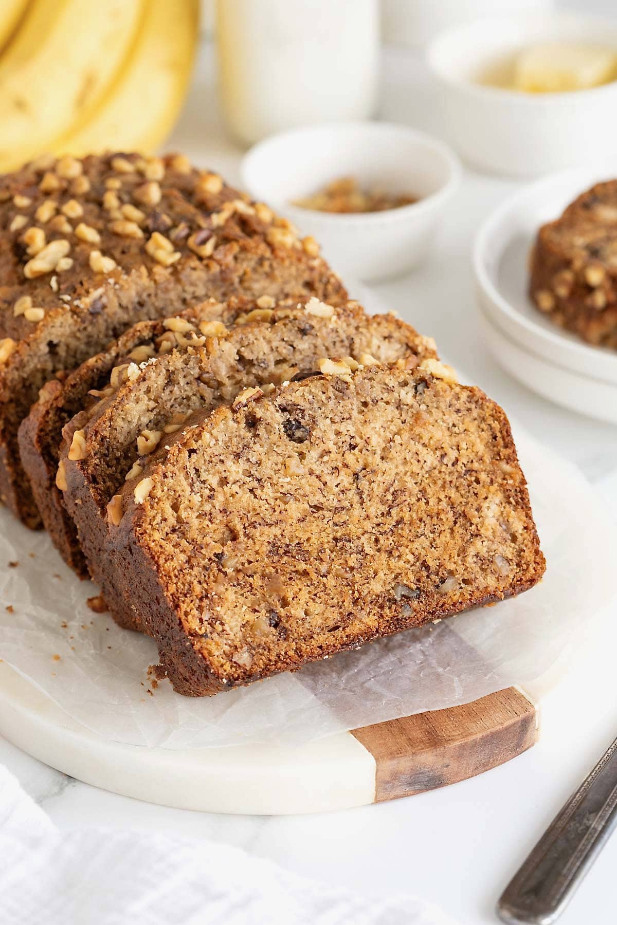 A loaf of banana nut bread on a white cutting board. Half of the loaf is cut into three thick slices.