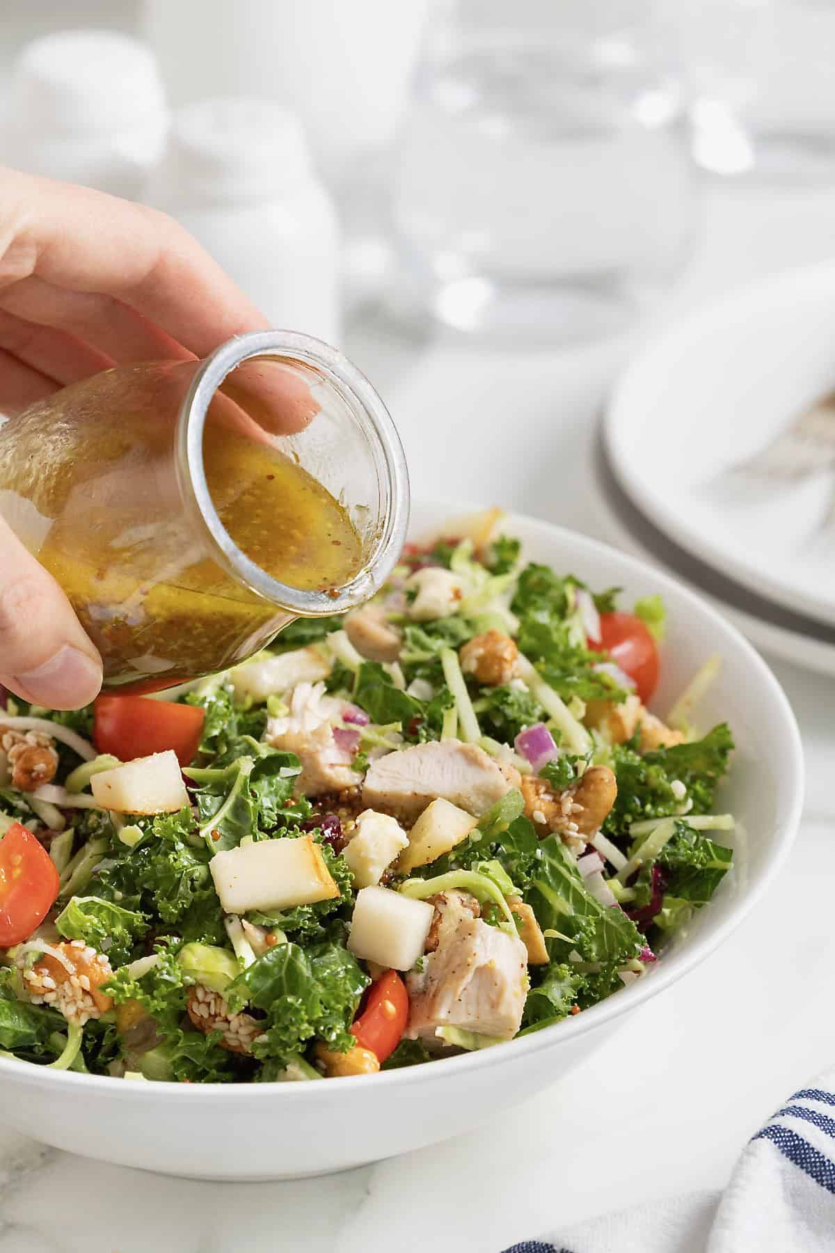 Champagne vinaigrette being poured from a small glass carafe onto a green salad with chicken and halved cherry tomatoes.