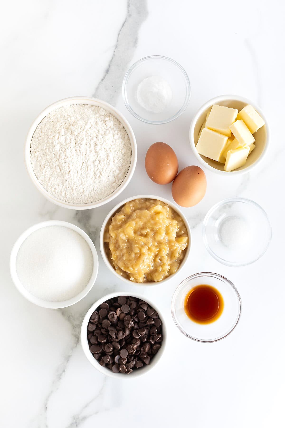Ingredients to make brown butter banana bread with chocolate chips in small white dishes on a white marble counter.